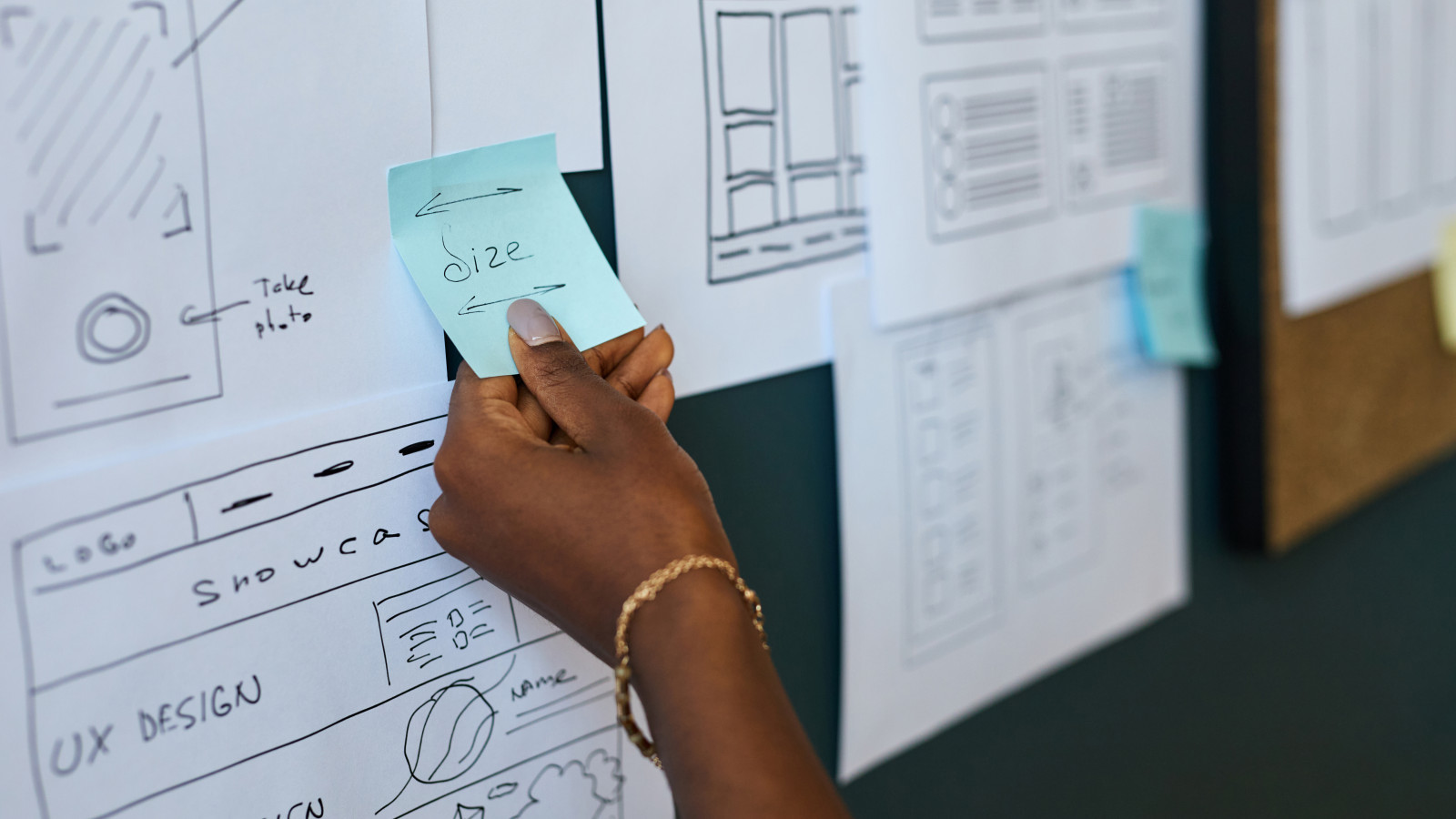 Black woman hand holding sticky note with word size written on it, placing note on wall covered with wireframe sketches and user interface design layouts during creative planning session